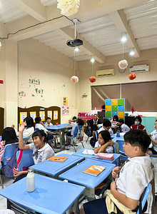 Students sitting on tables with one student raising her hand to ask the teacher a question.
