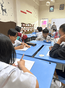 Group of students wearing white uniform in a circle writing on a table.