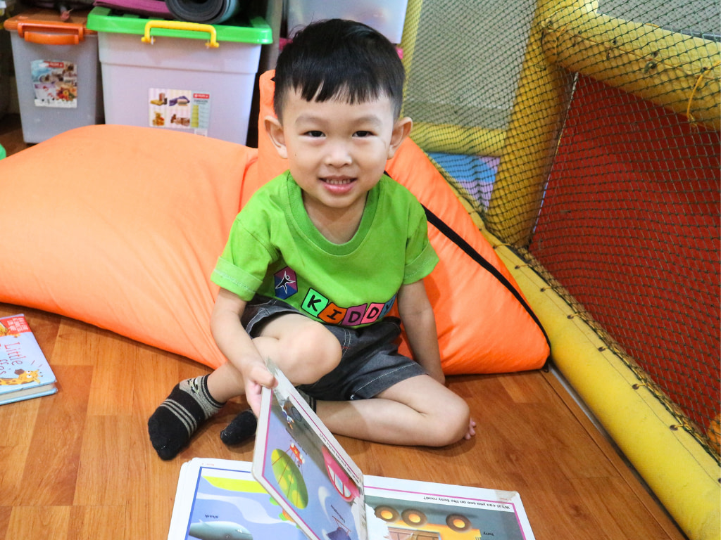 A playgroup student reading book while smiling on the wooden floor.