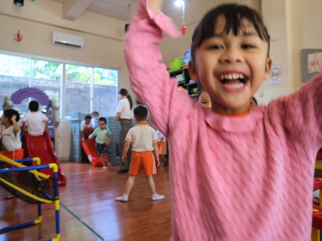 A young kindergarten student of Kiddy School Manado jumping and smiling while other students play in the background.