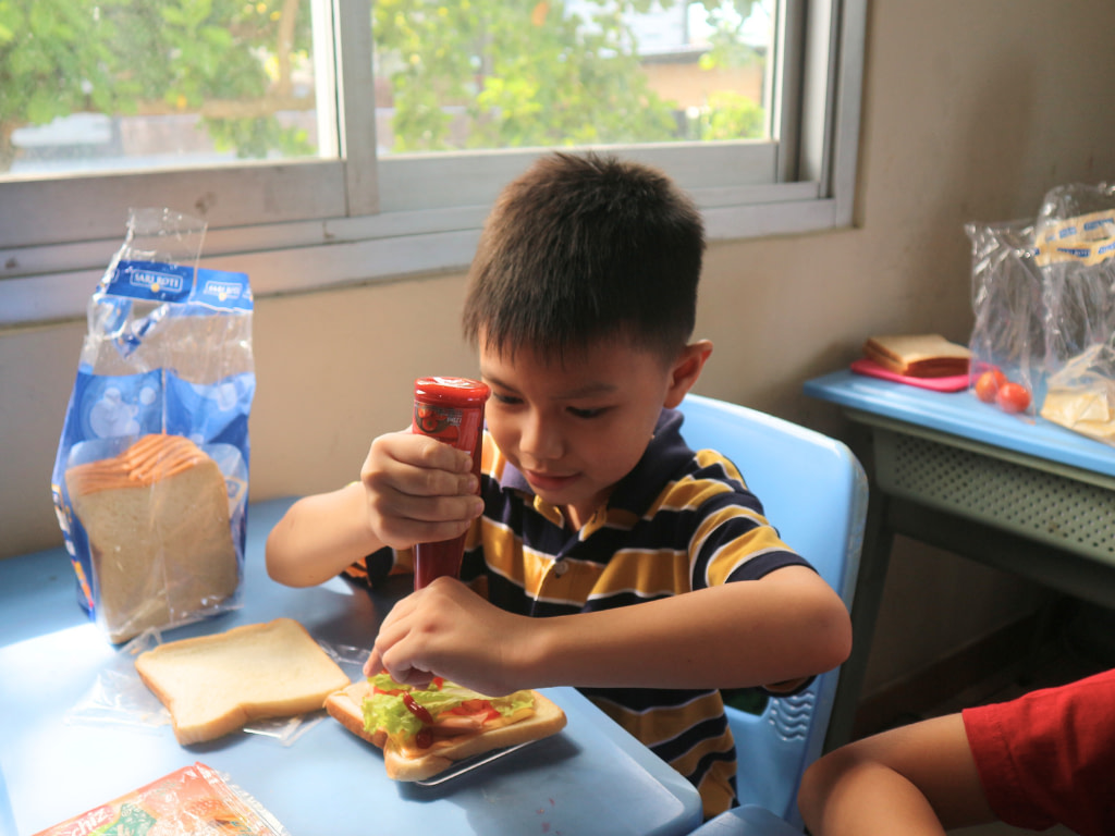 A young primary student sitting on a table and pouting ketchup on a sandwich in class.