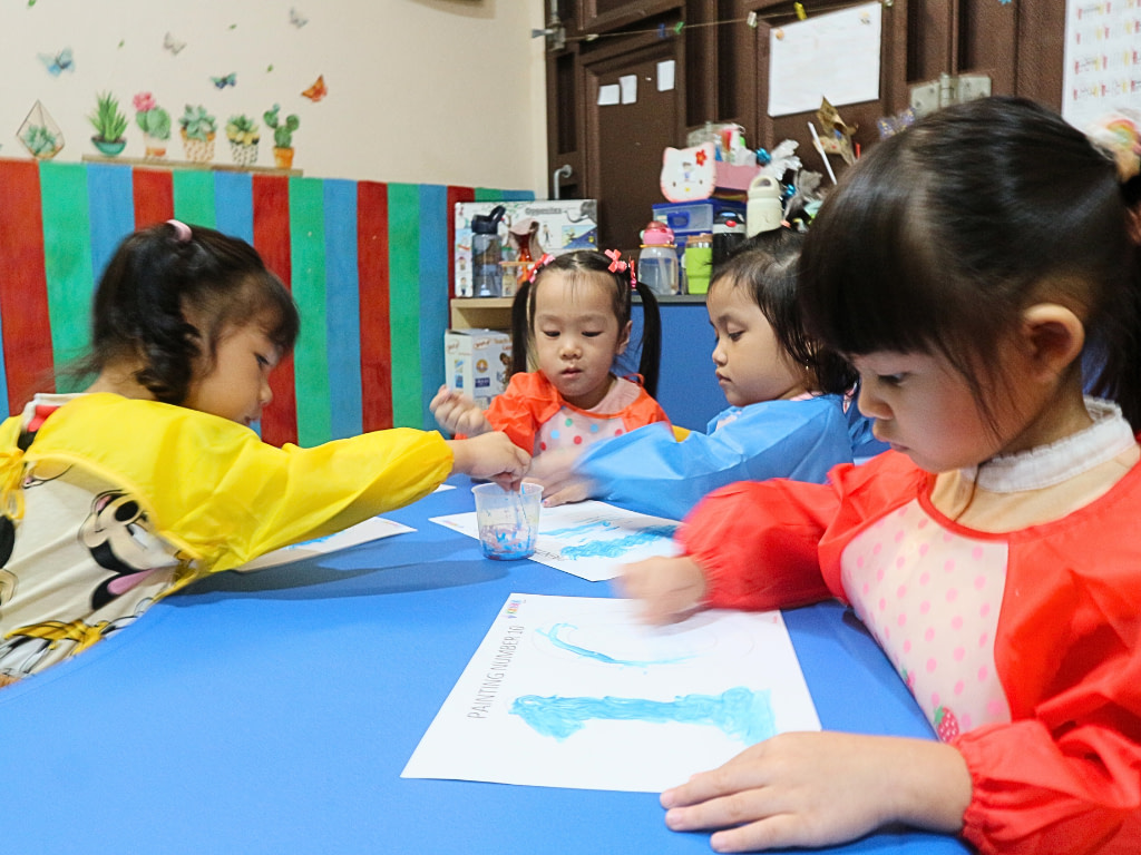 A group of playgroup students sitting on the table and painting while wearing colorful aprons.