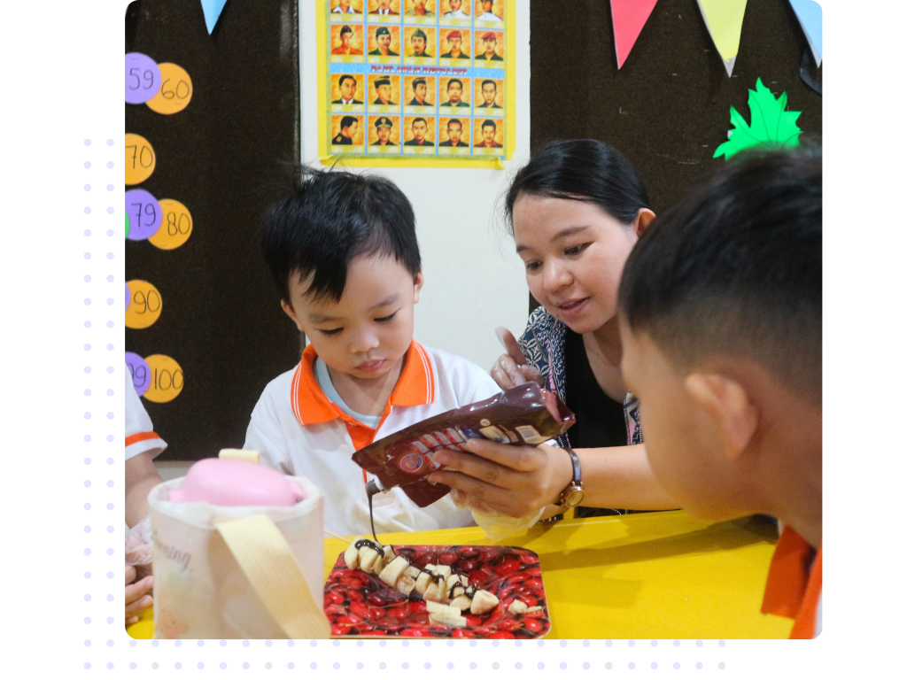 tk.3 A teacher guiding a student who is sitting and wearing a uniform while putting chocolate syrup on the plate.