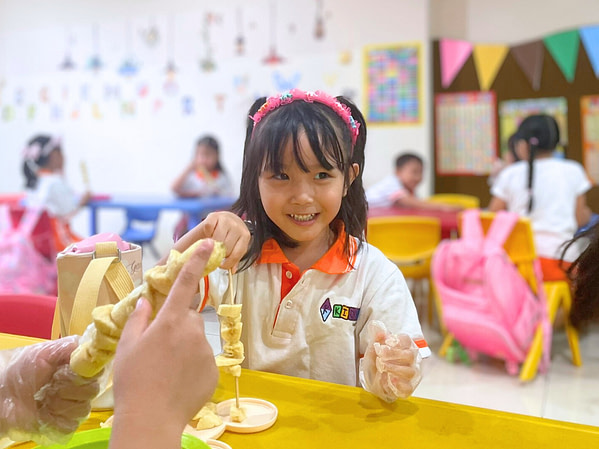 kindergarten.7 A young kindergarten girl smiling while holding a stick of banana with background of classroom filled with other students.